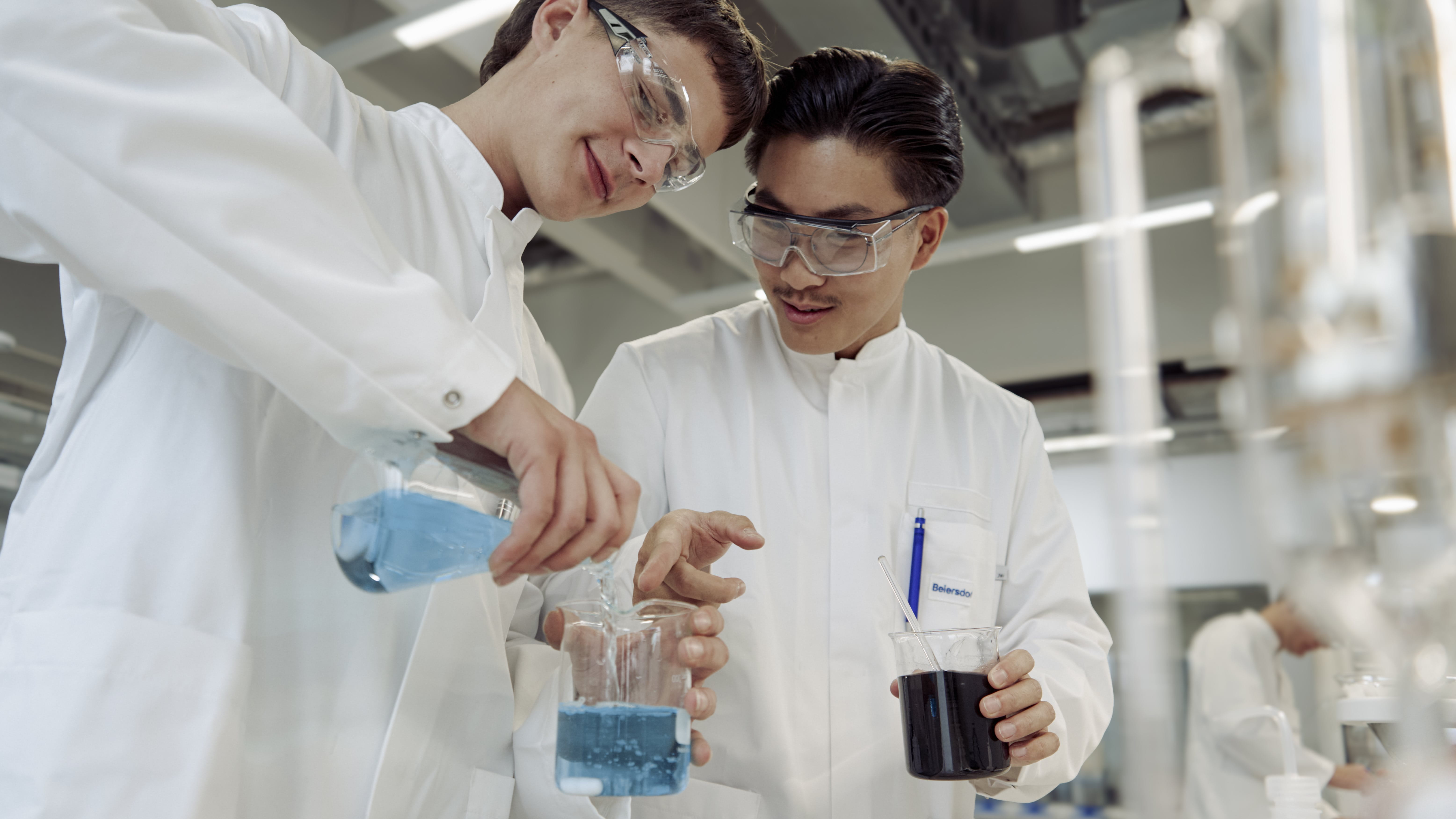 Two apprentices mix colorful liquids in the lab, wearing protective clothing and working together as a team.